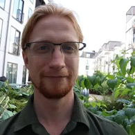 Foto von John Collins im Freien. Er hat lange rotblonde Haare und trägt eine Brille und ein grünes Hemd.