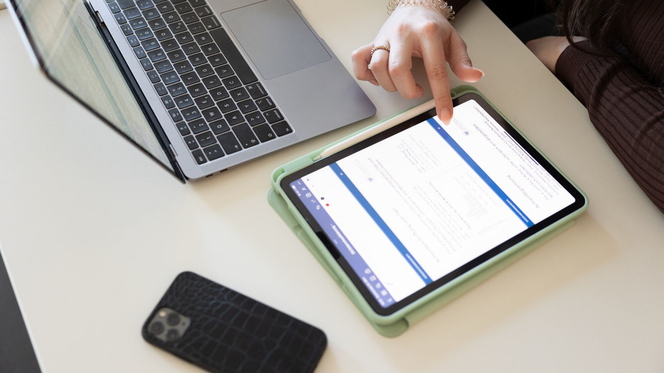 Eine Studentin tippt auf ihrem Tablet, ihr Handy und Laptop liegen daneben auf dem Tisch.