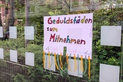 Bild von einem Zaun, auf dem ein Plakat hängt mit der Aufschrift "Geduldsfäden zum Mitnhemen". Darunter hängen Wollfäden.