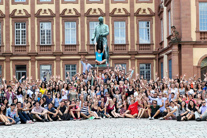 Eine große Gruppe an Studierenden sitzen im Ehrenhof vor einer Statue und winken.