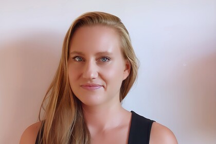 Katharina Ludwig hat lange blonde Haare und trägt ein schwarzes Tanktop. Sie steht vor einem weißen Hintergrund.