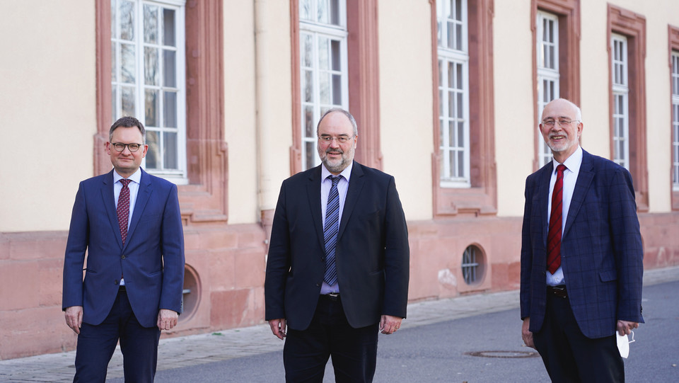 Drei Personen in anzug und Krawatte stehen vor dem Mannheimer Schloss. 