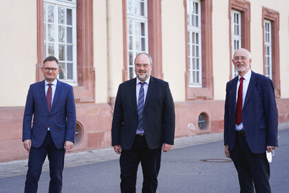 Drei Personen in anzug und Krawatte stehen vor dem Mannheimer Schloss. 
