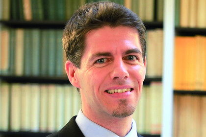 Portrait von Christoph Sajons. Er trägt ein hellblaues Hemd, sowie eine blau gestreifte Krawatte und einen schwarzen Blazer. Er steht lächelnd vor einem Regal.
