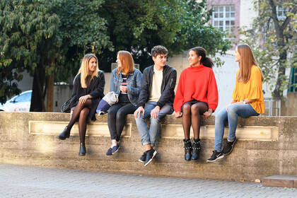 Fünf Studierende sitzen auf einer Mauer. Im Hintergrund Bäume und Unigebäude.