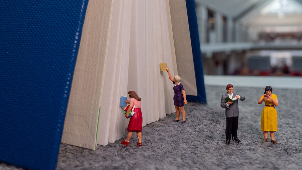 Zu erkennen ist ein großes Buch, zwei Miniaturfiguren lesen im Vordergrund, zwei weitere geben Bücher zurück oder lehen diese aus.