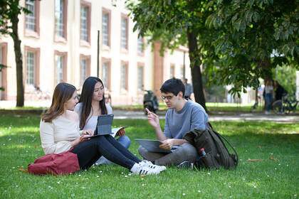 Drei Studierende sitzen auf dem Campus auf der Wiese. Sie haben Unterlagen und Laptops vor sich aufgeschlagen.