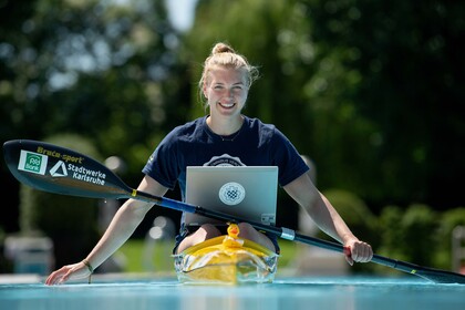 Sarah hat blonde Haare und trägt ein blaues Uni Mannheim T-Shirt. Sie sitzt in einem Kanu mit einer Quietscheente an der Spitze und einem Laptop auf ihrem Schoß. 