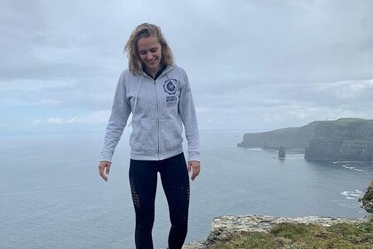 Muriel hat mittellange blonde Haare. Sie trägt eine graue Jacke. Im Hintergrund sind das Meer und Klippen zu sehen.  