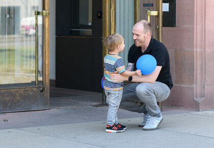 Eine erwachsene Person hockt vor dem Haupteingang zum Ostflügel der Universität Mannheim. Ein kleines Kind, das ihm gegenübersteht, schaut ihm direkt in die Augen und und hält dabei einen blauen Ballon. Beide scheinen in ein herzliches Gespräch vertieft zu sein.