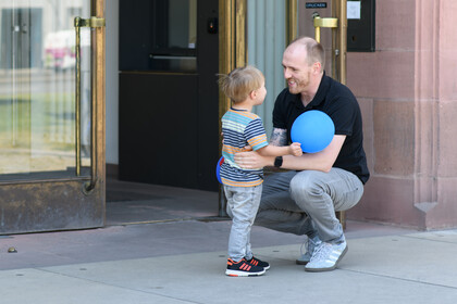 Eine erwachsene Person hockt vor dem Haupteingang zum Ostflügel der Universität Mannheim. Ein kleines Kind, das ihm gegenübersteht, schaut ihm direkt in die Augen und und hält dabei einen blauen Ballon. Beide scheinen in ein herzliches Gespräch vertieft zu sein.
