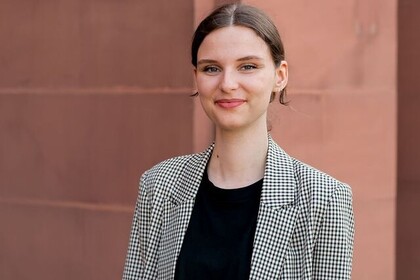 Portät von Bachelorstudentin Marie Abels. Sie trägt einen schwarz-weiß quarierten Blazer und ein schwarzes T-Shirt. Sie hat dunkelblonde Haare, die nach hinten gekämmt sind. Im Hintergrund ist eine rote Sandsteinmauer des Schlosses zu sehen. Link: Instagram-Post Portrait Marie Abels