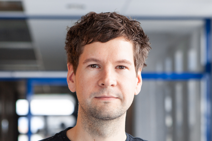 Christoph Kern in einem Flur mit Glastüren im Hintergrund. er trägt ein dunkles Shirt.