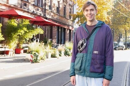 Porträt von VWL-Student Tim Bartelsen. Bartelsen trägt eine grün-violette Outdoorjacke, eine Gürteltasche über der Schulter und weite Jeans. Er steht auf den Schienen der Seckenheimer Landstraße in der Schwetzinger Vorstadt und lacht offenen in die Kamera. Link: Instagram-Post Stadtteile Mannheims