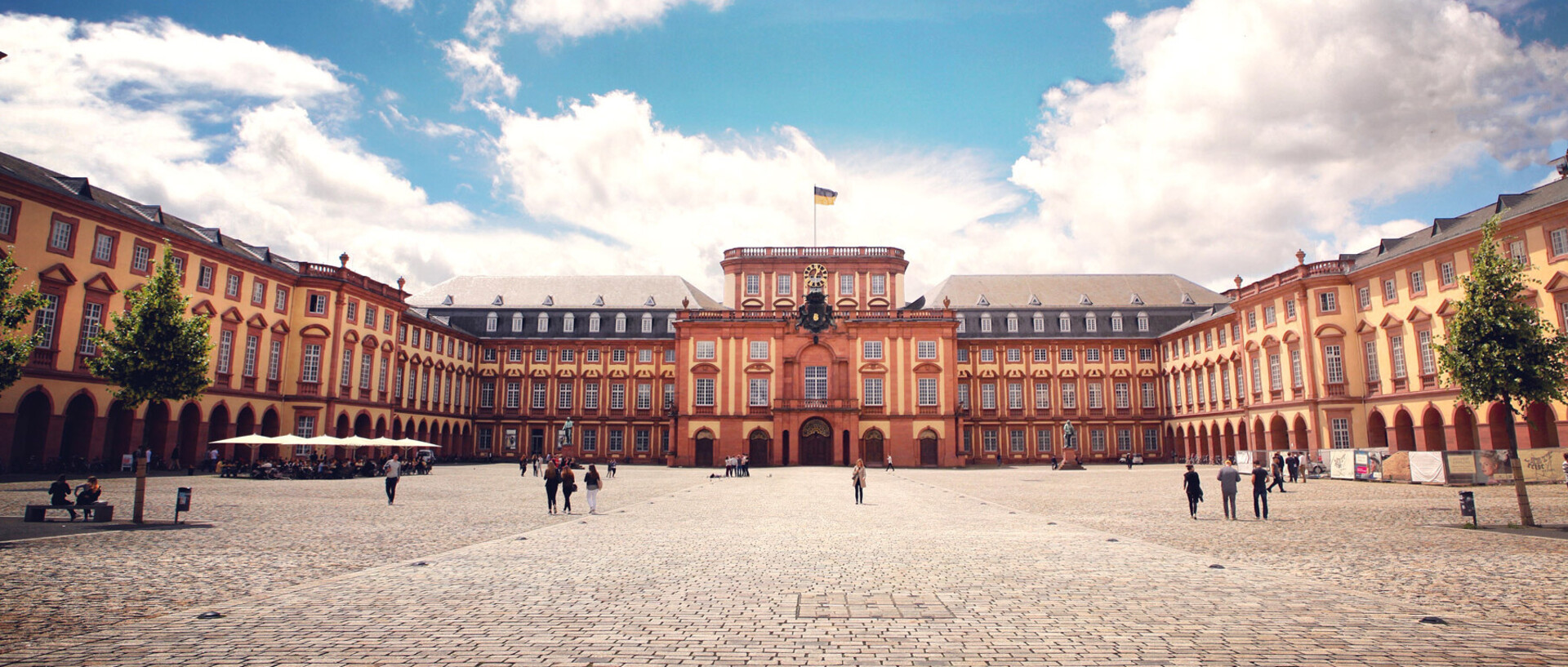 Das Barock-Schloss und der Ehrenhof der Universität Mannheim unter strahlend blauem Himmel. Das Schloss ist von unzähligen Fenstern, rotem Sandstein und einer gelben Fassade geprägt.