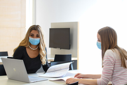 Zwei Frauen sitzen sich an einem Schreibtisch gegenüber. Beide tragen eine medizinische Maske und schauen auf einen Laptop.