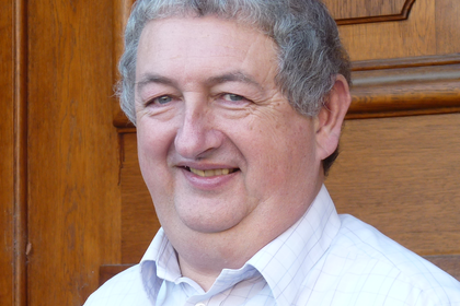 Portrait von Ian Bailey. Er trägt ein weißes Hemd und steht lächelnd vor einer braunen Holztüre.