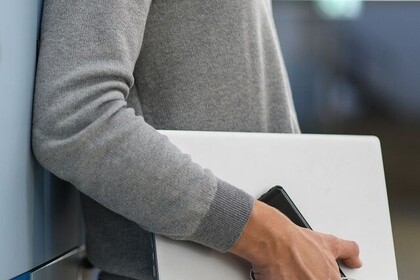 Ein Mann lehnt mit dem Rücken an einer hellblauen Wand. Er trägt einen grauen Pulli und einen Laptop unter dem Arm. Link: Instagram Post ENGAGE.EU