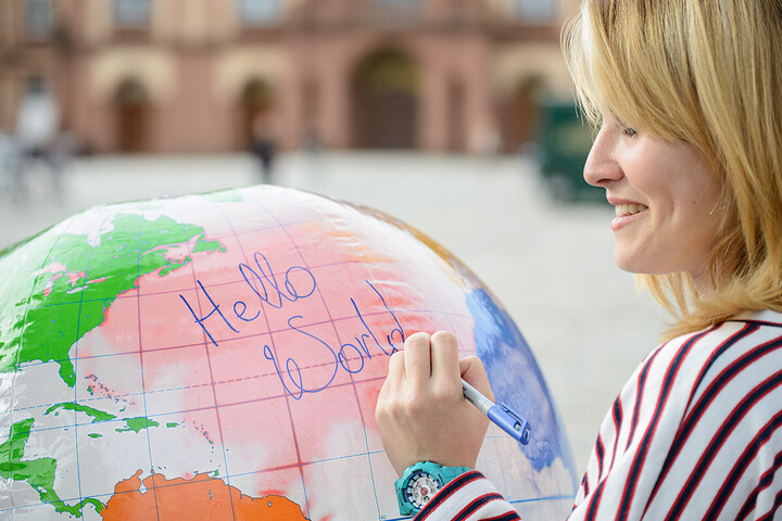 Eine blonde Frau schreibt mit einem blauen Stift auf eine Globus-Ballon „Hello World“. Sie steht auf dem Ehrenhof, hinter ihr das Mannheimer Schloss.