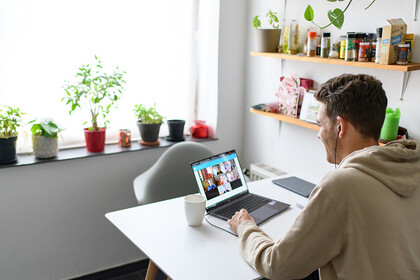 Studierender mit kurzen braunen Haaren und einem beigen Kapuzenpullover trägt Kopfhörer und sitzt an einem Tisch. Vor ihm steht ein Laptop, auf dem ein Online-Meeting mit mehreren Personen zu sehen ist.