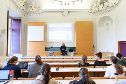 In einem Hörsaal steht eine Dozentin am Rednerpult und hält eine Vorlesung. In den Stuhlreihen davor sitzen Studierende und hören zu. Der Raum ist modern ausgestattet. Nur an der Decke sind Stuckverzierungen zu erkennen. Link: Instagram-Post Treffen der internationalen Universitätsallianz U7+
