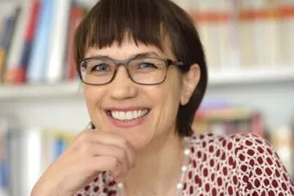 Portrait von Annette Kehnel. Sie trägt eine rot-weiße Bluse und sitzt lächelnd vor einem Bücherregal.