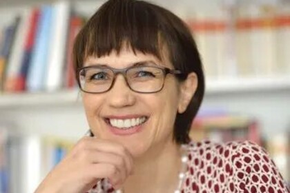 Portrait von Annette Kehnel. Sie trägt eine rot-weiße Bluse und sitzt lächelnd vor einem Bücherregal.