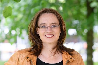 Portrait von Michele Tertilt. Sie trägt einen schwarzen Pollover sowie eine orange farbige Jacke und steht auf einer Grünfäche.