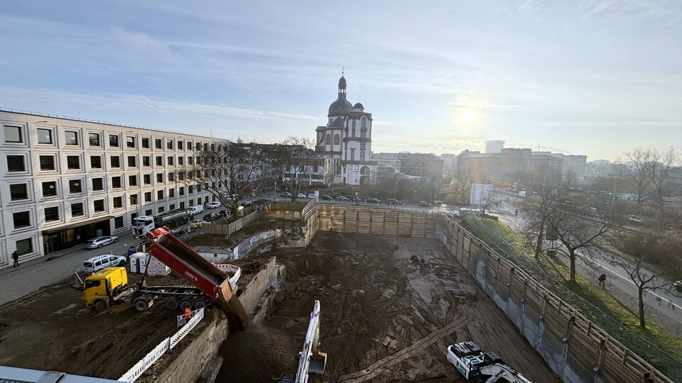 Im Vordergrund die Baustelle in A5 mit großer Baugrube, im Hintergrund die Jesuitenkirche Mannheim bei Sonnenaufgang. Ein Lkw kippt Erde in die Grube, daneben zwei Bagger. Links das modernes Gebäude B6, rechts Bäume und eine Straße.