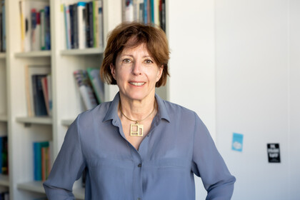 Eine lächelnde Person trägt eine blaue Bluse und steht vor einem Bücherregal. Die Person heißt Michaela Wänke.
