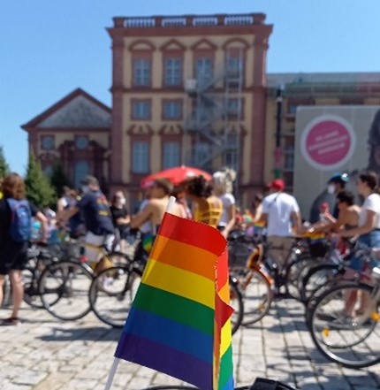 Fahrrad mit Regenbogen-Fahne im Vordergrund und im HIntergrund viele Menschen auf Fahrrädern.