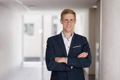 Jens Förderer. He is wearing a black suit and is standing inside a white walled building.