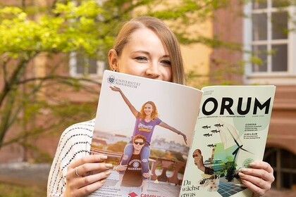 Eine weiblich gelesene Studierende liest vor dem Hauptgebäude in dem Universitätsmagazin "Forum". Sie hält das Heft hoch zu ihrem Gesicht und schaut über den Rand in die Kamera.