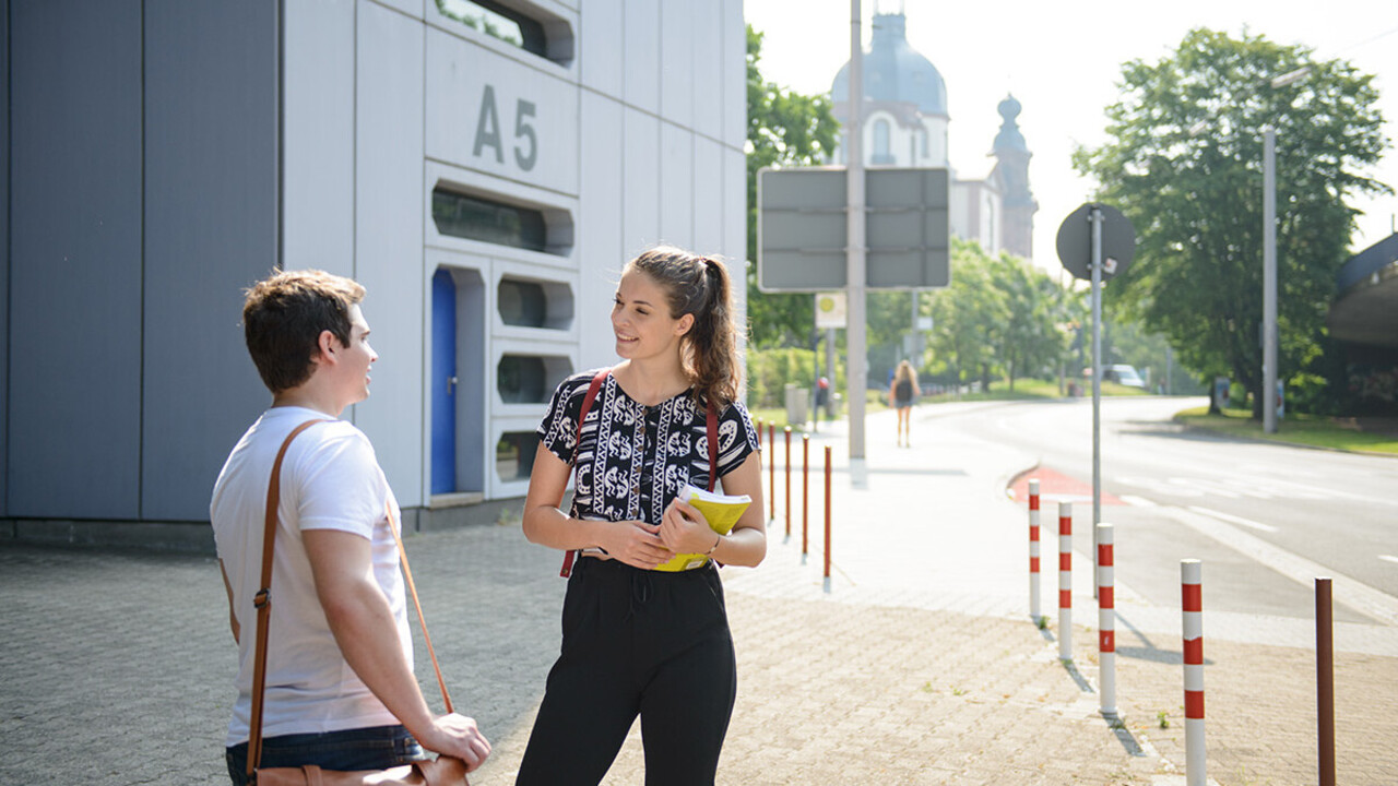 Zwei Studierende stehen gemeinsam vor dem A5-Gebäude an der Straße und unterhalten sich.