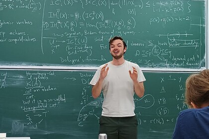 Junger Mann steht vor einer Tafel und spricht in Richtung einer Klasse