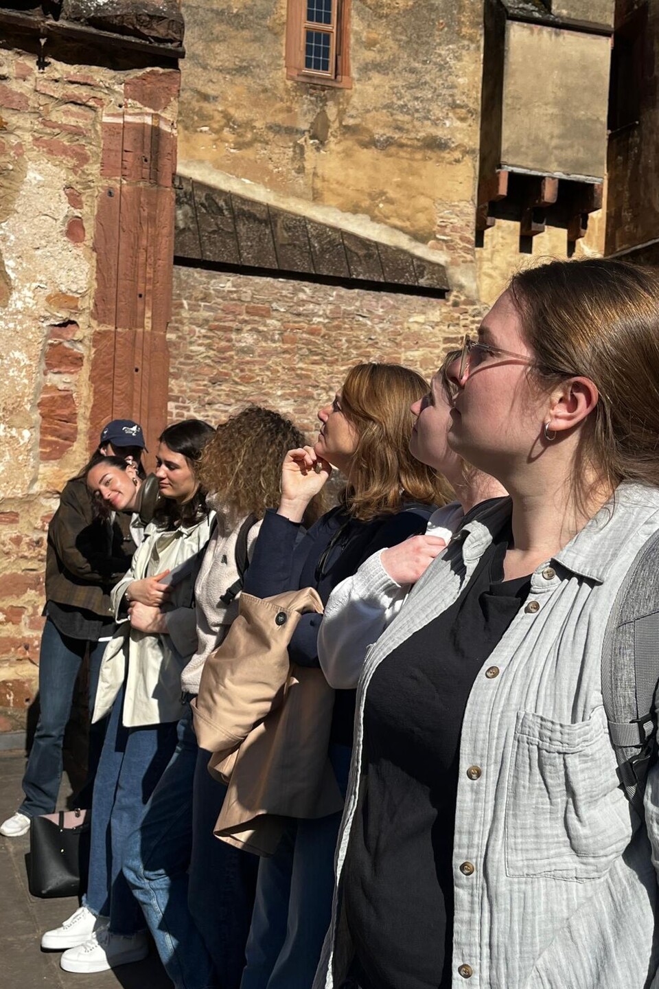 7 nach links blickende Frauen vor einem historischen Gemäuer.