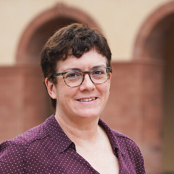 Jane Ossandon-Schönfeld hat braune Haare und eine Kurzhaarfrisur. Sie trägt eine lila Bluse mit weißen Punkten und eine Brille.