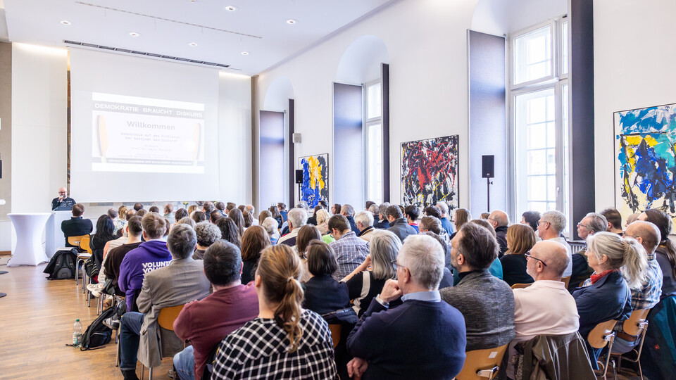 Viele Zuschauer im Fuchs-Saal hören dem Redner am Rednerpult zu. Foto von Hinten aufgenommen