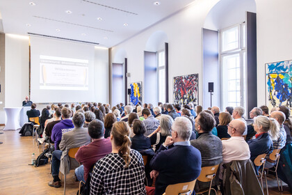 Viele Zuschauer im Fuchs-Saal hören dem Redner am Rednerpult zu. Foto von Hinten aufgenommen