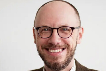 Portrait von Gerhard Ernst. Er trägt eine Brille, ein blau-weiß kariertes Hemd und ein braunes Sakko.