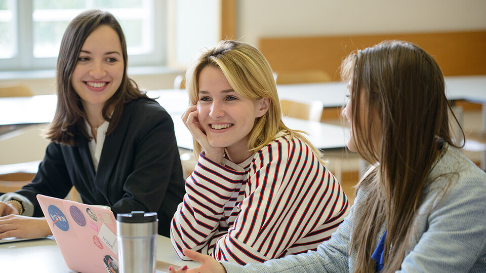 Drei Studentinnen sitzen in einem Seminarraum und lachen. Vor ihnen steht ein aufgeklappter Laptop. Die beiden äußeren Frauen schauen beide zur mittleren.