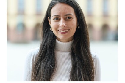 Porträt der Studentin Magdalena Tsureva in der Reihe #testimonialtuesday. Sie lächeltoffen und frpöhlich in die Kamera. Tsureva trägt einen weißen Rollkragenpullover und große Ohrringe und ihre lange schwarze Haare offen. Link: Instagram-Story Testimonial Tuesday