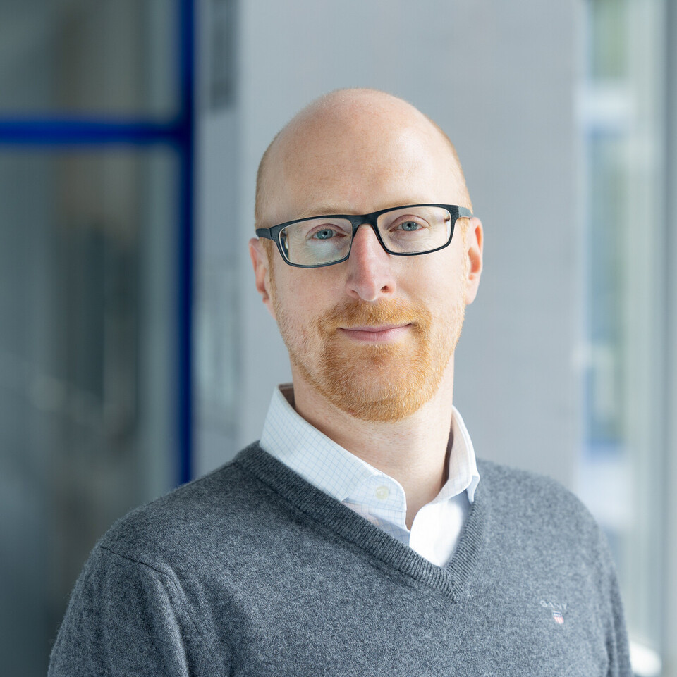 Florian Keusch trägt ein weißes Hemd und darüber einen grauen Pullover.