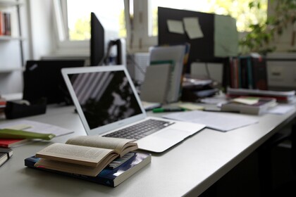 Büro mit aufgeklapptem Laptop und aufgeschlagenem Buch auf Schreibtisch