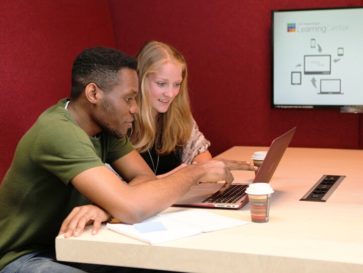 Zwei gutgelaunte Studenten sitzen in Bibliothek an einem Tisch und schauen auf gemeinsamen Laptop. Sie zeigen mit ihren Fingern auf den Bildschirm.