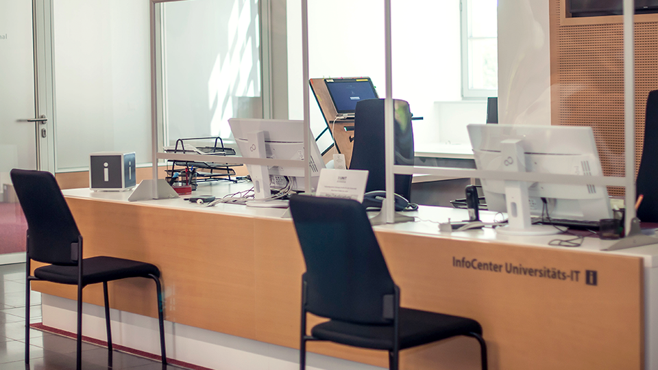 Im Bild befindet sich die Infotheke der Universitäts-IT. Auf der einen Seite sind zwei Stühle für Mitarbeitende vor Bildschirmen und auf der anderen Seite befinden sich zwei Stühle für Besucher*innen.