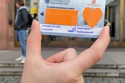 Eine Hand hält die ecUM-Karte der Uni Mannheim in die Kamera. Namen und Porträt der Karteninhaberin sind mit orangenem Papier überklebt. Link: Instagram Post Infos zur ecUM-Karte