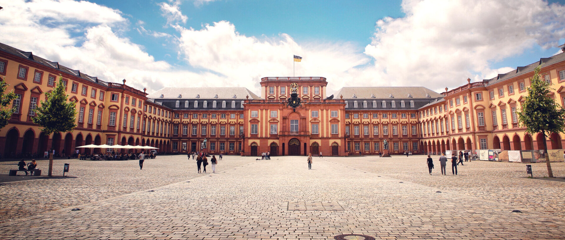 Das Barock-Schloss der Universität Mannheim steht unter blauem Himmel. Es ist von unzähligen Fenstern, rotem Sandstein und einer gelben Fassade geprägt.