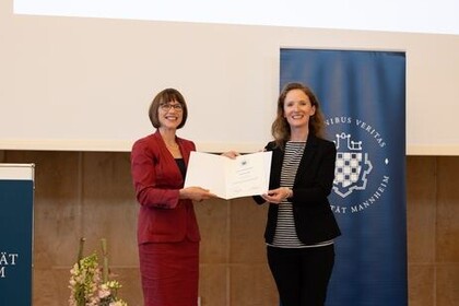 Prof. Dr. Karin Hoisl (rechts, in dunklem Kostüm) erhält den Lehrpreis der Universität Mannheim und hält diesen gemeinsam mit der verleihenden Person (links, rotes Kostüm) in die Kamera.