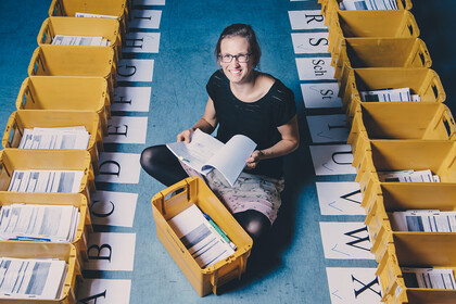 Eine Mitarbeitende der Universität Mannheim sitzt auf dem Boden. Um sie herum stehen gelbe Postkisten. Die Kisten sind mit verschiedenen Buchstaben beschriftet.